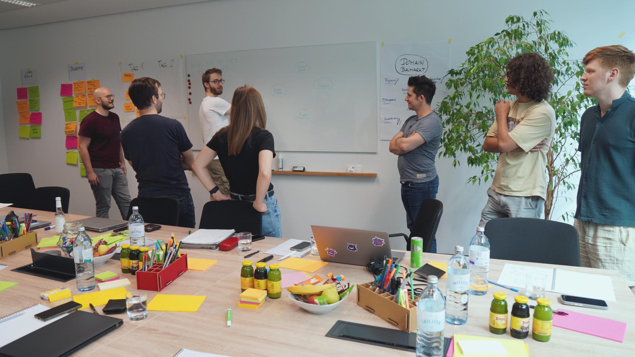 A group of people standing in front of a whiteboard, one is drawing. In the forground, a table with sticky notes, pens, laptops and more stuff.