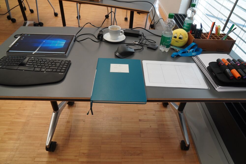 Teacher's table with laptop in tablet/drawing configuration, keyboard, mouse, a paper notebook, coffee, a soft ball, flip chart markers and more